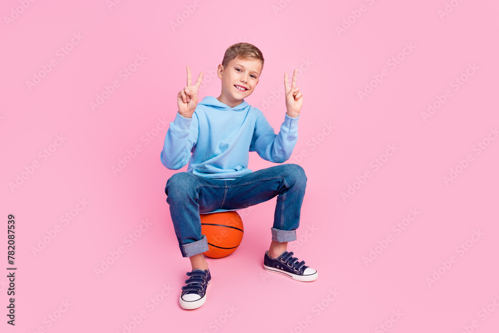 Fototapeta premium Full size photo of little cheerful boy sit basketball show v-sign empty space isolated on pink color background