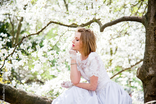 beautiful blonde smiling romantic bride in a white dress sitting on a branch in blossoming magnolia garden on sunny spring day