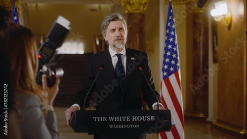US presidential candidate pronounces inspirational political speech. Minister or congressman speaks at press conference in the White House, answers journalists questions. American flags in background.