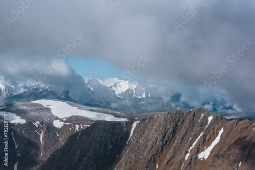 Dramatic aerial top view to rocky mountain range and large snowy mountain peak in low clouds. Atmospheric alpine landscape with sheer crags and snow-capped top in low cloudiness. Big snow mountains.