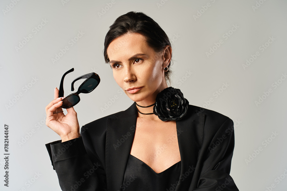 A fashionable woman in a sleek black suit confidently holds up a pair of chic sunglasses, exuding a sense of sophistication and style. - obrazy, fototapety, plakaty 