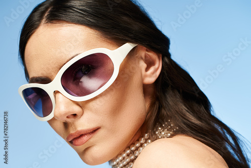 A stylish woman with a serene expression wearing trendy white sunglasses in a studio setting with a vibrant blue background.