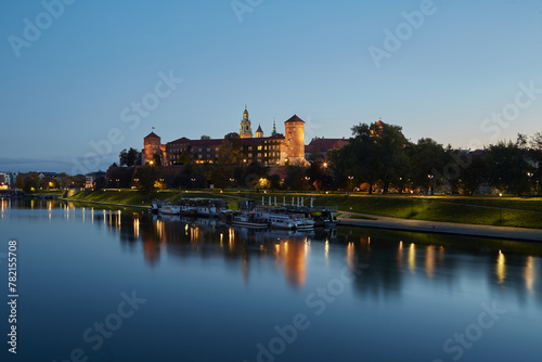 Krakow, Poland. 09.10.2022 Historic Wawel Castle in the evening blue hour on the banks of the Vistula river and floating ships restaurants.