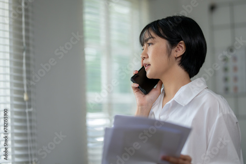 Free Download Serious young woman in white shirt speaking on mobile phone while holding papers, in a bright office. - GFXLoad