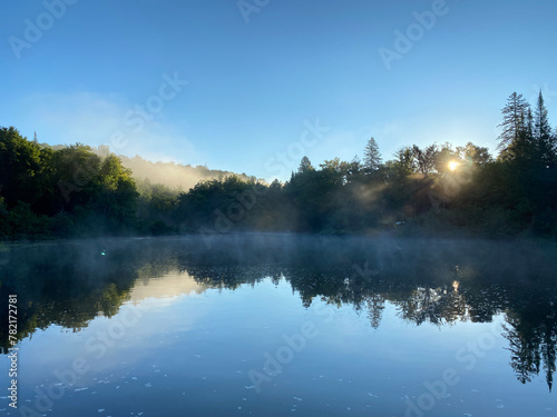 Morning on the river. Quiet forest and river at dawn. Reflection of a mountain on a lake in the early twilight. Sunrise with mist on the water. Sunbeams through the fog.