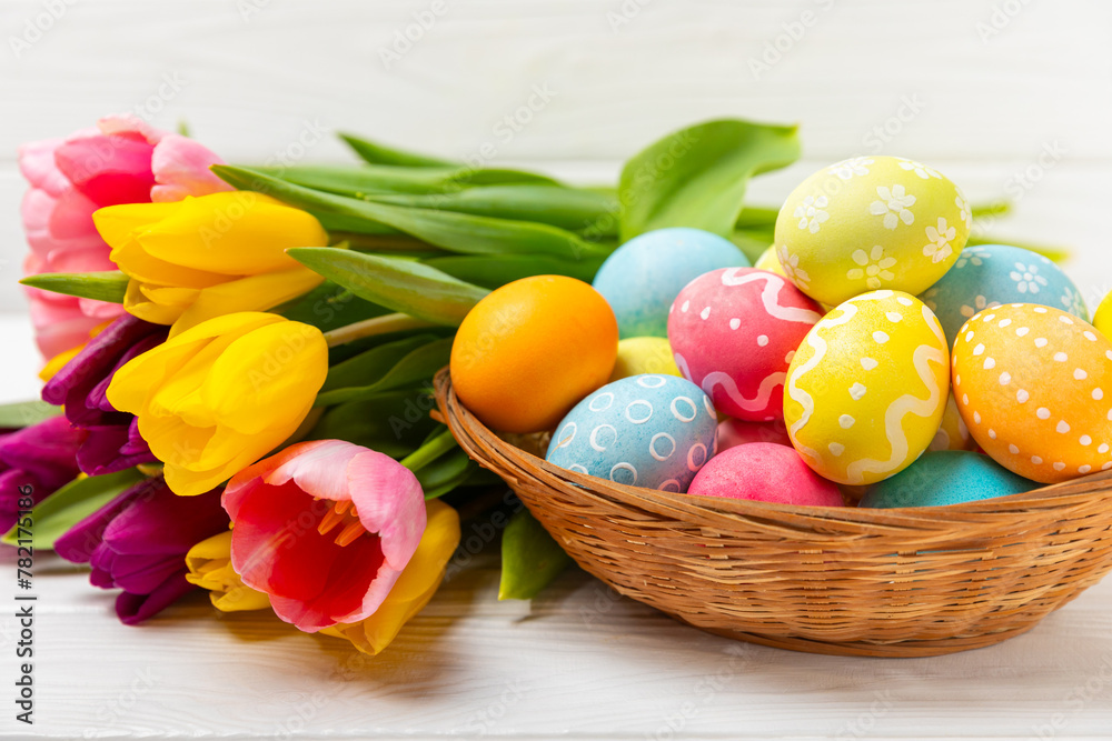 Easter basket filled with colorful eggs and a bouquet of tulips on a textured wooden table. Easter celebration concept. Colorful easter handmade decorated Easter eggs. Place for text. Copy space Easter basket filled with colorful eggs and a bouquet of tulips on a textured wooden table. Easter celebration concept. Colorful easter handmade decorated Easter eggs. Place for text. Copy space