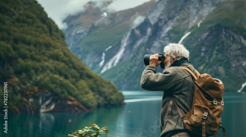 Senior hiker observing mountainous landscape through binoculars by the lake