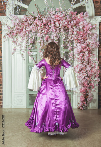 Young beautiful woman in fantasy rococo style medieval dress standing near wall with pink flowers back pose