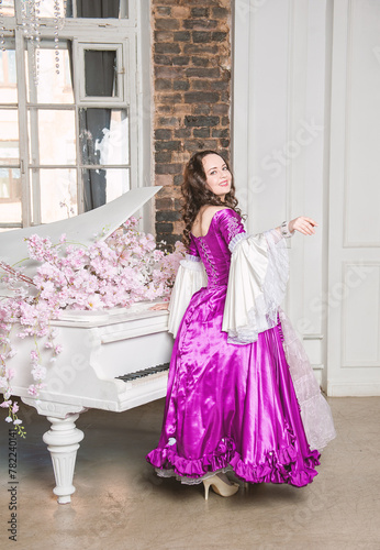 Young smiling beautiful woman in fantasy rococo style medieval dress standing turn near piano with pink flowers