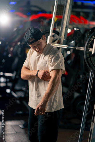 in the gym young handsome pumped up guy in a T-shirt and cap stands looking at his biceps