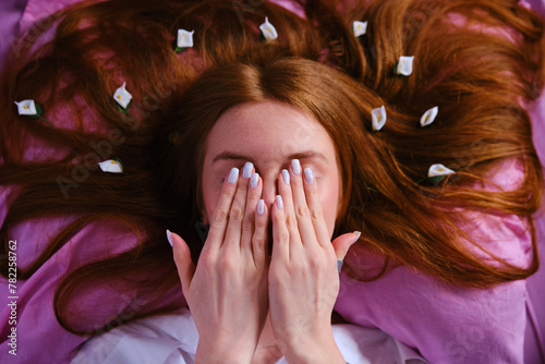 Young redhead Teenager Girl with blue eyes and white flowers in red hairs Relaxing On pink Bed At Home Wearing white shirt and closing his eyes with hands playing peekaboo