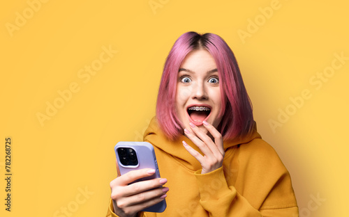Shocked astonished very happy pink woman wear braces, orange hoodie open mouth, use hold hand cell phone, modern smartphone, mobile phone, isolated yellow wall background. Online offer, dental care ad