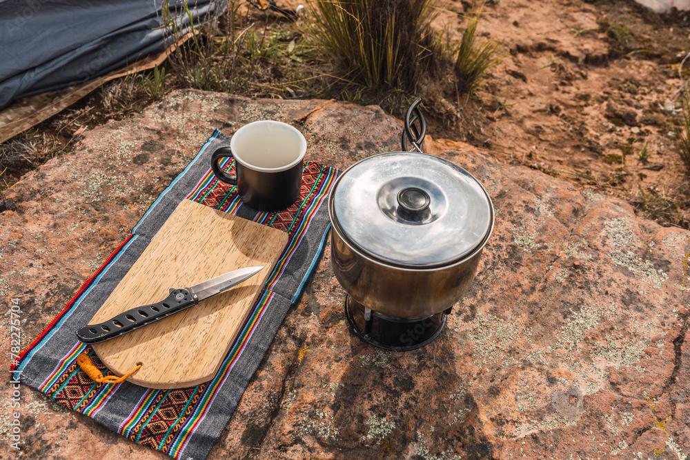 set of camping utensils pots knife knife cup and chopping board placed on the vegetation in the Andes mountain range with a lake background and blue sky with clouds set of camping utensils pots knife knife cup and chopping board placed on the vegetation in the Andes mountain range with a lake background and blue sky with clouds