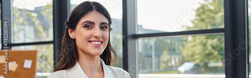 Free Download A businesswoman stands in front of a window, smiling brightly as sunlight illuminates her face, embodying a sense of empowerment and positivity. - GFXLoad