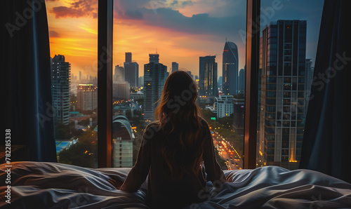 back view of young woman wake up at luxury hotel room or apartment, person by panoramic window, city and skyscrapers photo