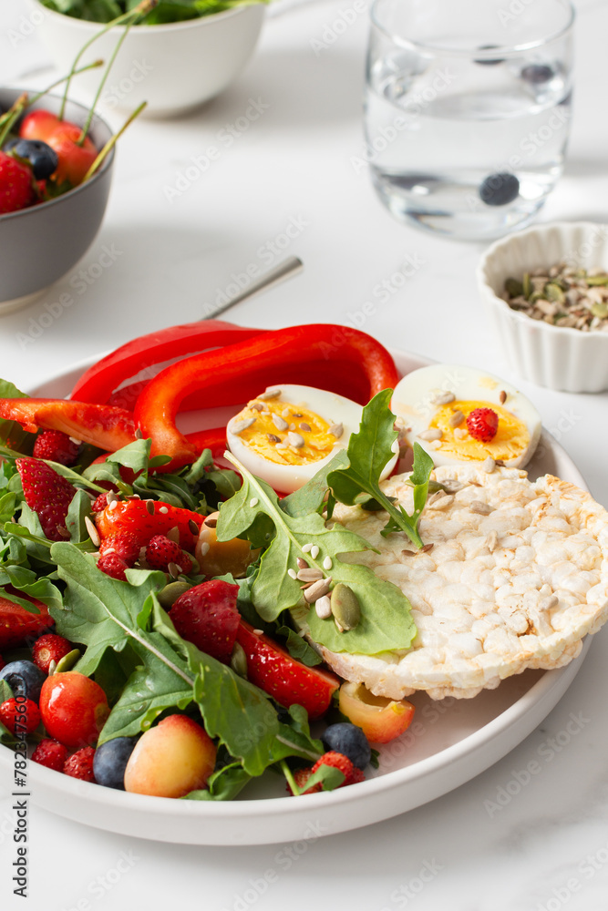 Healthy tasty breakfasts, a plate with boiled eggs and rice bread, salad of arugula and strawberries with cherries and blueberries, fresh peppers and seeds