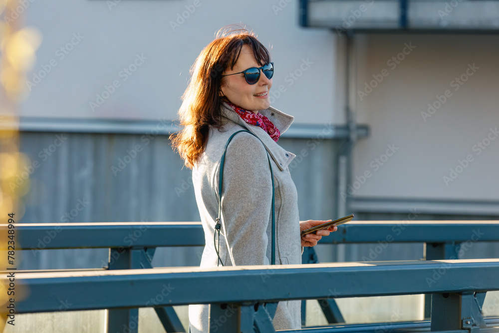 Fototapeta premium Smiling woman enjoying a sunny morning in the city, dressed in casual chic attire with a vibrant scarf and smartphone in hand, concept: leisure, urban lifestyle, relaxation