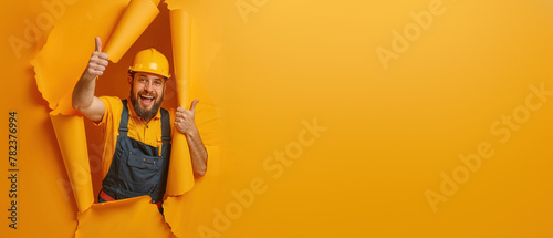 A bearded man in a hard hat with a smile poses through a hole in a yellow background