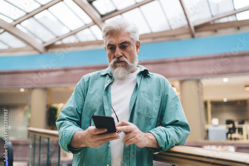 Concentrated elderly Caucasian man with beard using smartphone app in bright office. The senior, in his 60s, seems absorbed by cellular, reflecting tech-savvy generation against indoor modern backdrop