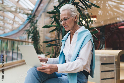 Elegant senior woman with silver hair, using a laptop, focused and engaged, seated in a bright mall, technology literacy in the elderly. Mature businesswoman using laptop computer in offce
