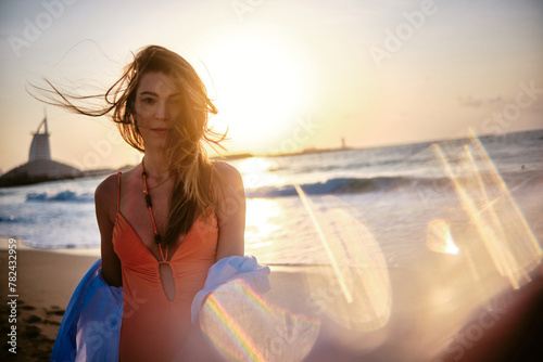 A woman stands on a beach holding a wind-blown blanket, with a beautiful sunset and a distant lighthouse in the background