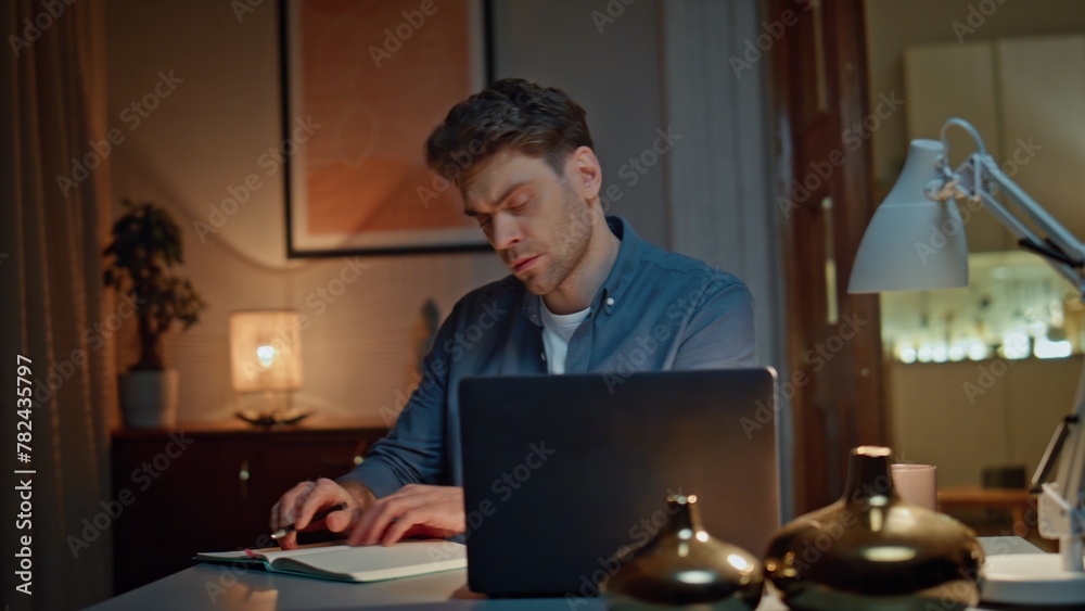 Serious guy writing notes watching laptop at night home closeup. Freelance man Serious guy writing notes watching laptop at night home closeup. Freelance man