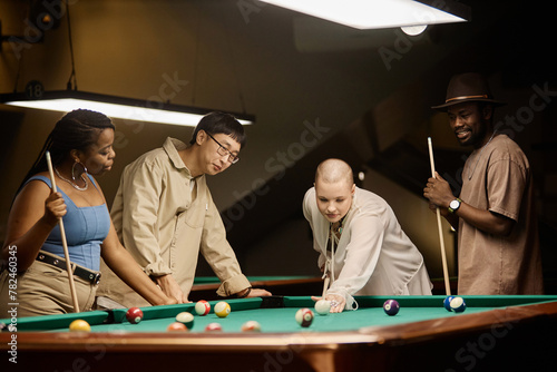 Portrait of multiethnic group of friends playing pool together by table in nightclub in muted tones