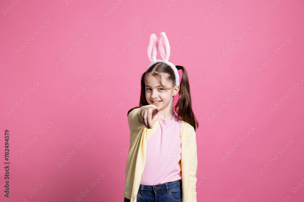Young schoolgirl with pigtails dancing around in pink studio, pointing at something in front of the camera and having fun. Joyful small child feeling confident with bunny ears for easter.
