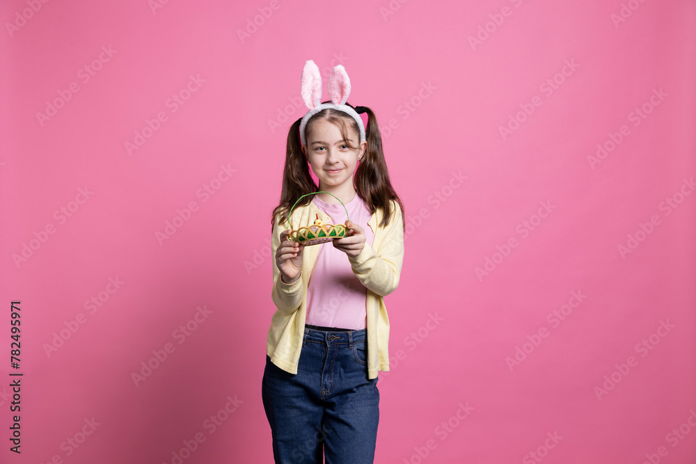 Adorable little girl showing a basket filled with easter eggs and a chick, posing with confidence in front of camera. Cheerful small child holding spring decorations, pigtails and bunny ears.