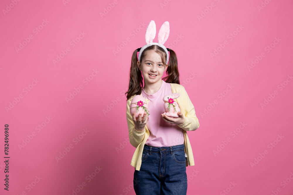 Young cute kid with bunny ears showing pink easter ornaments in front of camera, holding her handmade egg and rabbit toy. Small child smiling in studio and celebrating spring holiday.