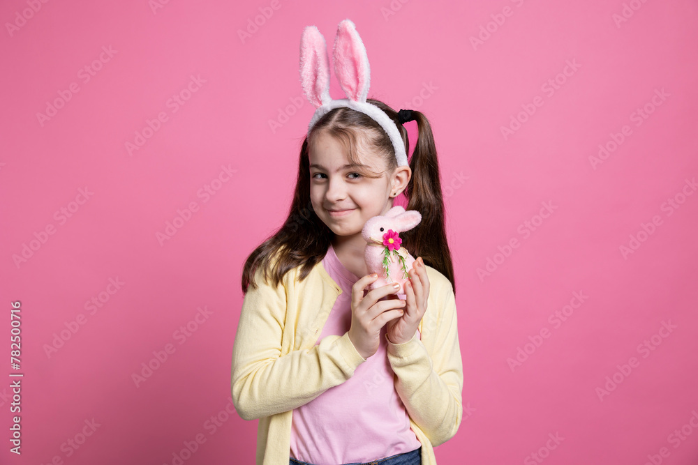 Positive cheerful girl with bunny ears posing over pink background, feeling enthusiastic about easter festive celebration. Cute small child holding a pink rabbit toy in front of camera, fluffy items. Positive cheerful girl with bunny ears posing over pink background, feeling enthusiastic about easter festive celebration. Cute small child holding a pink rabbit toy in front of camera, fluffy items.