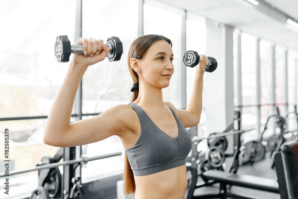 young athletic caucasian woman trains in fitness gym, standing and doing dumbbell press up, brunette girl in grey top and leggings, healthy lifestyle concept young athletic caucasian woman trains in fitness gym, standing and doing dumbbell press up, brunette girl in grey top and leggings, healthy lifestyle concept
