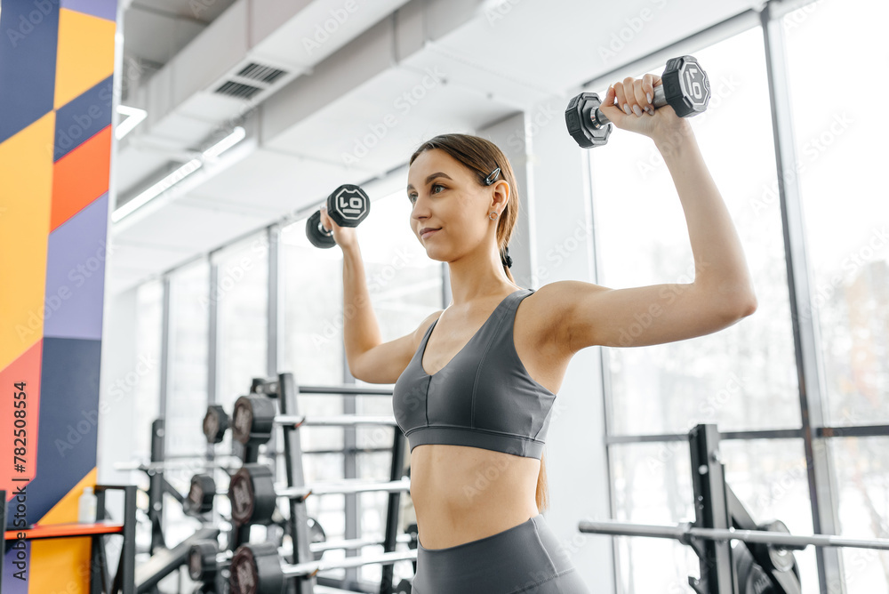 young athletic caucasian woman trains in fitness gym, standing and doing dumbbell press up, brunette girl in grey top and leggings, healthy lifestyle concept young athletic caucasian woman trains in fitness gym, standing and doing dumbbell press up, brunette girl in grey top and leggings, healthy lifestyle concept