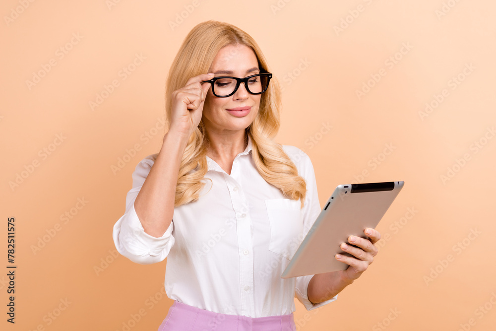Portrait of confident business person mature age serious woman focused reading results using tablet isolated on beige color background