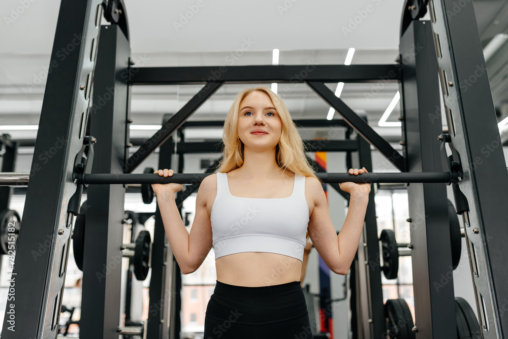 young athletic caucasian woman trains in fitness gym, squats with barbell in smith simulator, blond girl in white top and black leggings, healthy lifestyle concept young athletic caucasian woman trains in fitness gym, squats with barbell in smith simulator, blond girl in white top and black leggings, healthy lifestyle concept