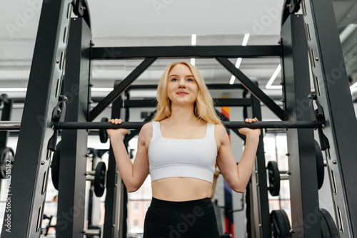 young athletic caucasian woman trains in fitness gym, squats with barbell in smith simulator, blond girl in white top and black leggings, healthy lifestyle concept