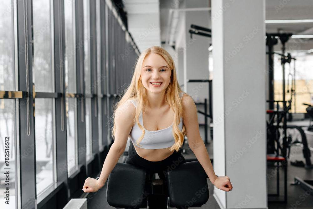 Fototapeta premium young athletic caucasian woman trains in fitness gym, ready to do hyperextension on exercise equipment and smiling, blond girl in white top and black leggings, healthy lifestyle concept