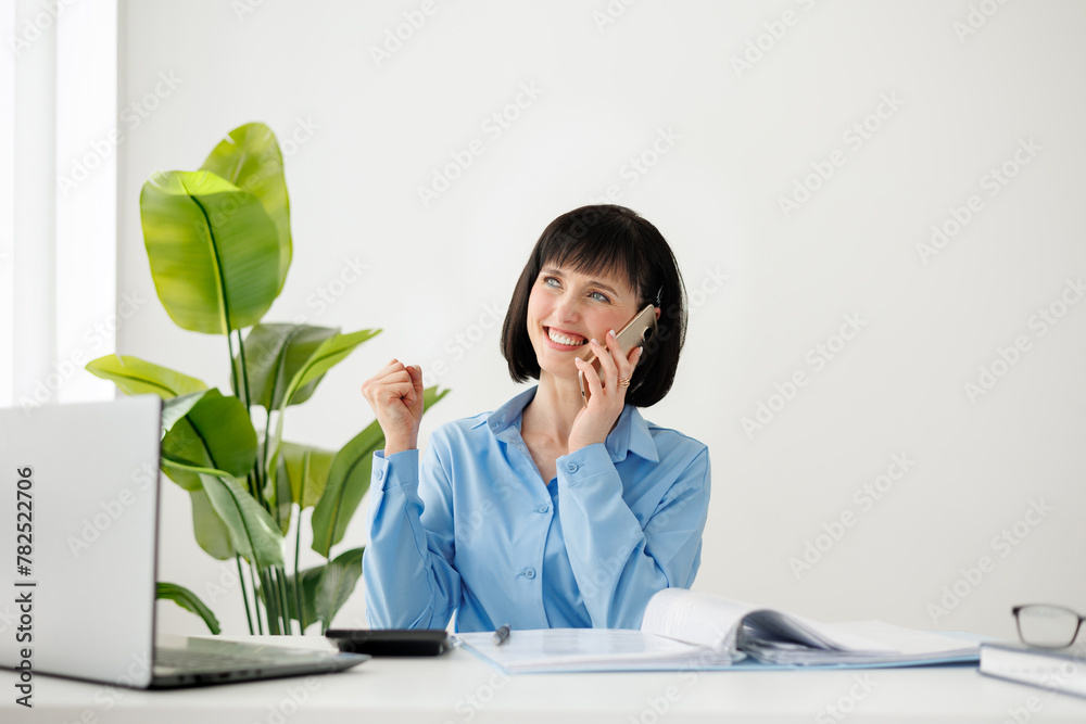 Excited successful employee businesswoman talking on smartphone say yes doing winner gesture, clench fist, sit at office desk with laptop. Achievement career, making a deal, business success concept