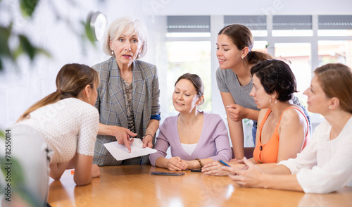 during meeting,senior employee stands at table holding documents in hands and argues her position. Using printed report, young student explains to colleagues how to achieve goal