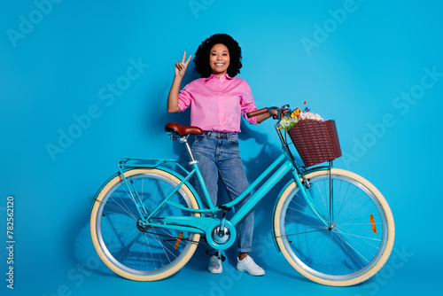 Full size photo of cheerful nice woman wear pink shirt hold bicycle showing v-sign say hi greetings isolated on blue color background © deagreez