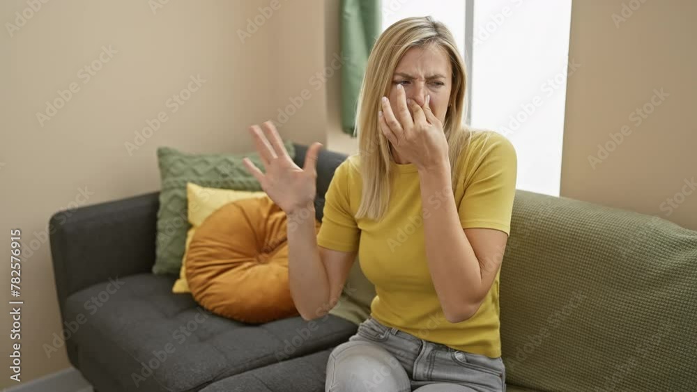 Young, attractive blonde woman dons t-shirt indoors, grimacing as disgusting, intolerable stench invades her home. nose pinched, holding breath to block smelly stink. disgust visible. Young, attractive blonde woman dons t-shirt indoors, grimacing as disgusting, intolerable stench invades her home. nose pinched, holding breath to block smelly stink. disgust visible.