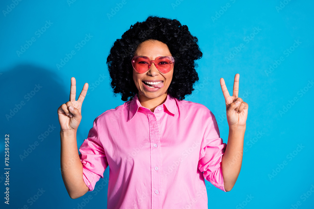 Portrait of pretty young woman show tongue out v-sign wear pink shirt isolated on blue color background Portrait of pretty young woman show tongue out v-sign wear pink shirt isolated on blue color background