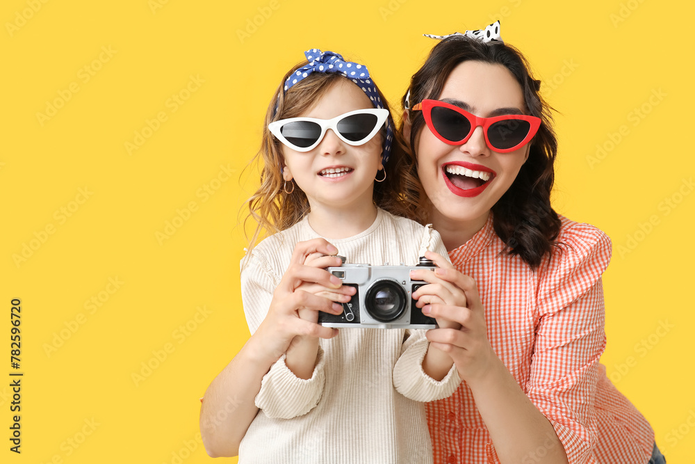 Fototapeta premium Beautiful pin-up woman and her daughter with photo camera on yellow background