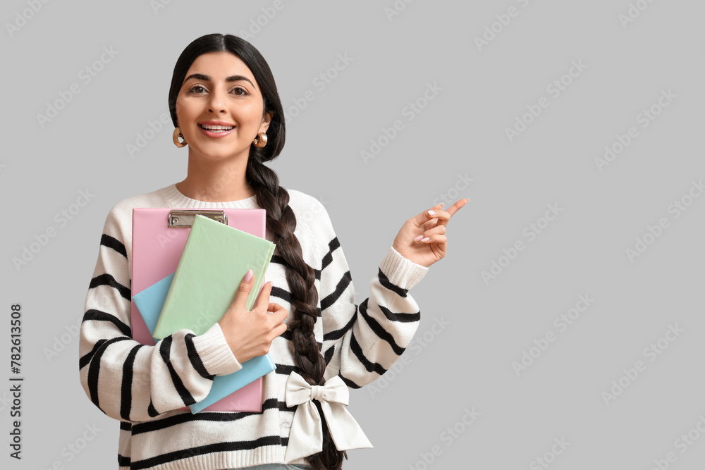 Beautiful young happy Indian woman with notebooks pointing at something on grey background - obrazy, fototapety, plakaty 