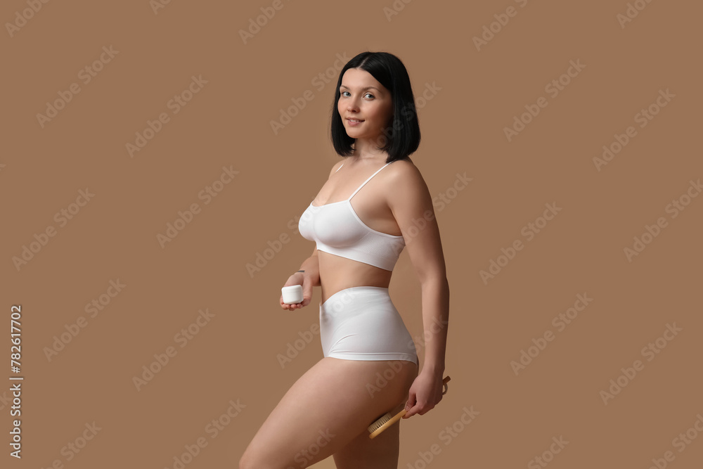 Beautiful young happy woman with massage brush and jar of anti-cellulite cream on brown background Beautiful young happy woman with massage brush and jar of anti-cellulite cream on brown background