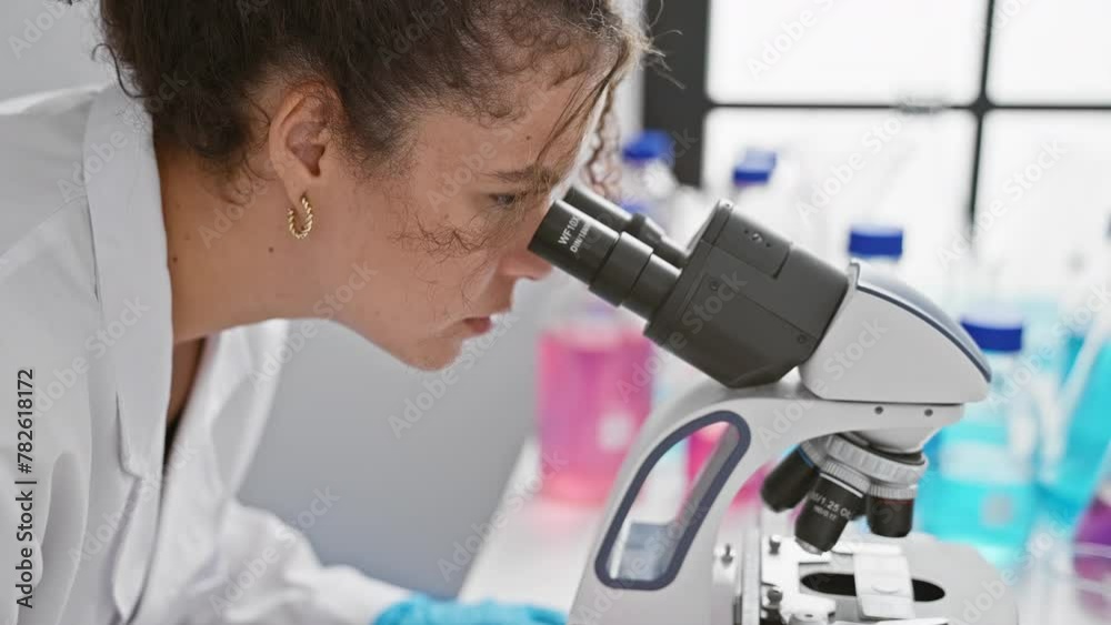 A young hispanic woman with curly hair examines samples using a microscope at a laboratory, symbolizing research and scientific discovery. A young hispanic woman with curly hair examines samples using a microscope at a laboratory, symbolizing research and scientific discovery.