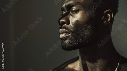 Against a backdrop of dark shadows a confident black man stands tall with his head held high. The interplay of light and shadows on his chiseled features highlights his refined sense .