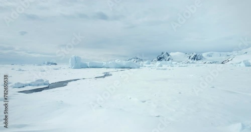 Fly over snow hill in Antarctica winter landscape. Frozen polar ocean with melted cracked river. Glacier and iceberg stuck in ice coast bay. Mountain range in background. Arctic travel and exploration