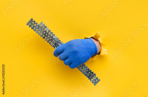 A left man's hand in a blue knitted glove holds a U-shaped fastening profile for drywall. Torn hole in yellow paper. The concept of a worker, a labor migrant, a master of his craft. Copy space.