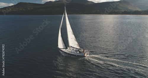 Luxury yacht at open sea racing under sails aerial. Amazing seascape of ocean bay with ship. Epic sail boat at Brodick pier harbor, Arran island, Scotland, Europe. Cinematic summer cruise